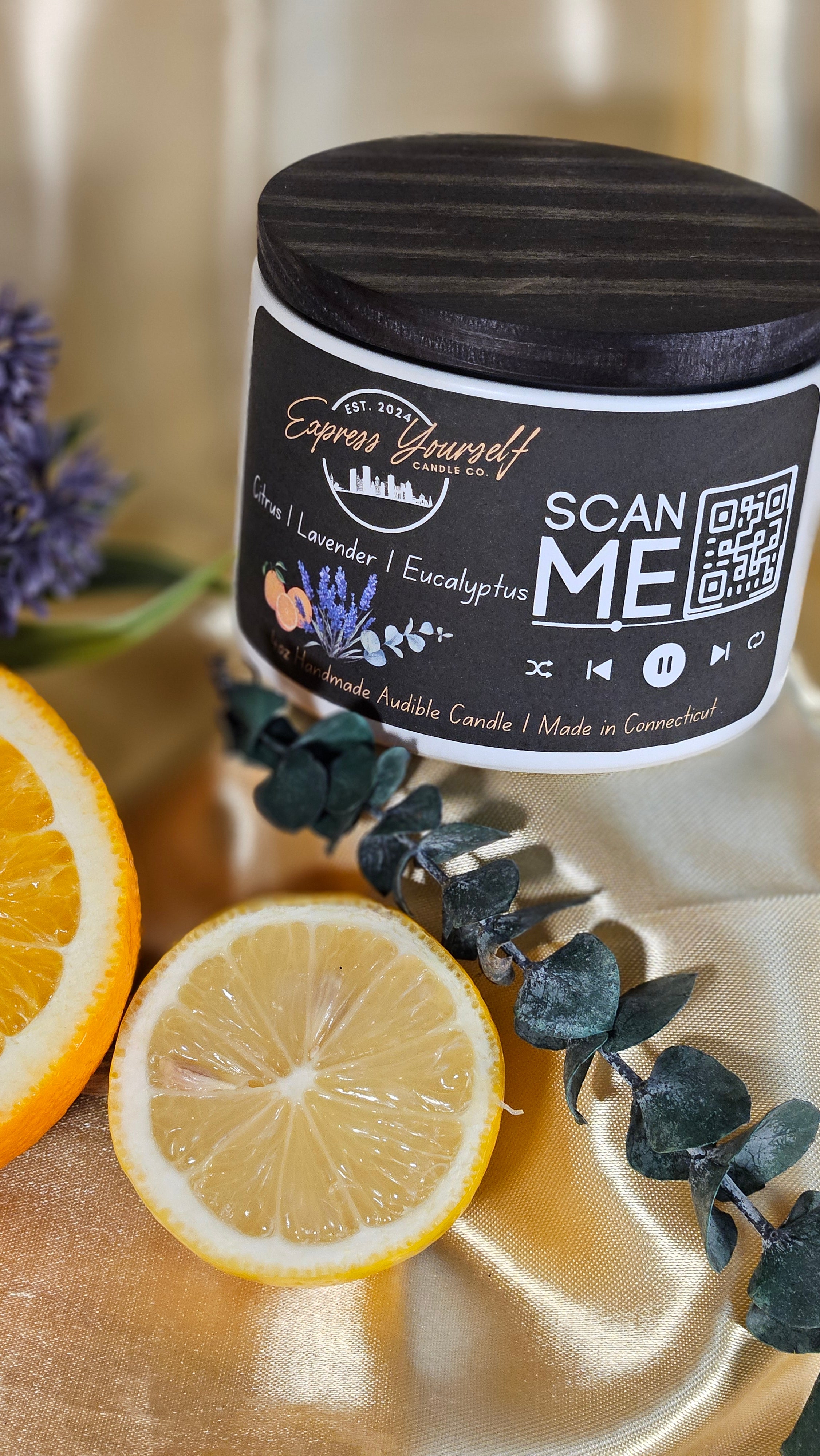 A scented mini messenger candle with a label that reads 'Express Yourself Candle Co.', placed on a gold surface with a decorative eucalyptus branch and a slice of lemon next to it.